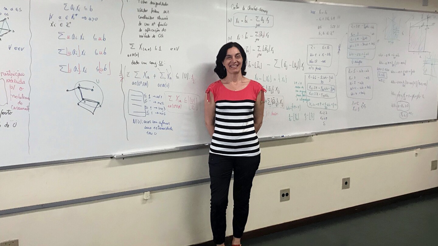 Luciana Buriol is seen standing in front of a large whiteboard filled with equations, she is standing toward the center, smiling with her hands clasped behind her back