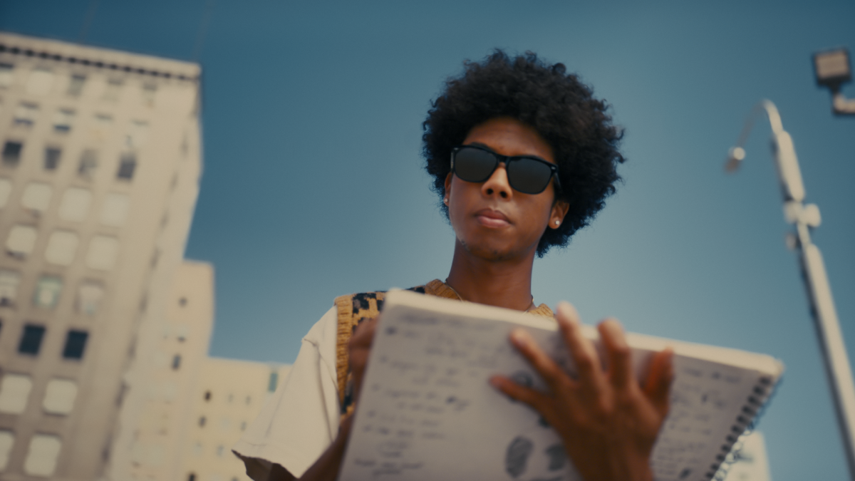 A person is seen wearing Echo Frames sunglasses outside. The person carries a notebook and is looking down at it, and there are some buildings and blue sky in the background.