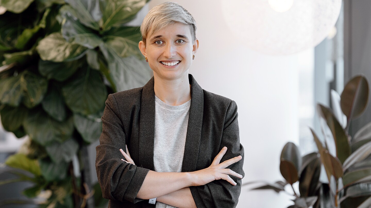 Olga Moskvyak, an applied scientist, is seen smiling into the camera, her arms are crossed and there are some plants in the background