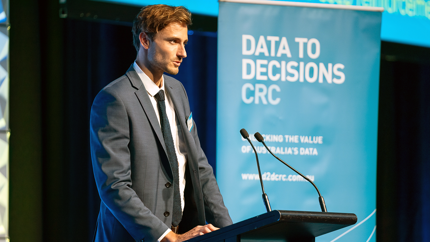 Alexander Long is seen wearing a suit, speaking at podium, the banner behind him and to the right says data to decisions CRC
