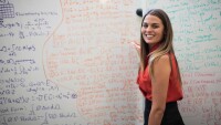 Danielle Maddix Robinson is seen standing in front of a whiteboard covered in calculations, she is looking over her left shoulder and smiling at the camera