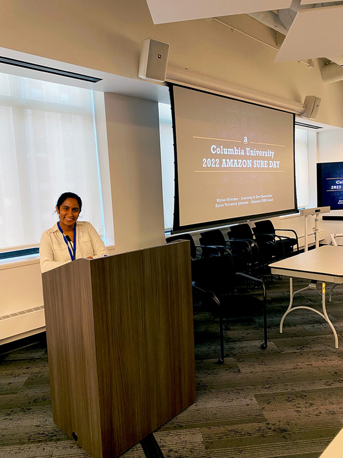 Maryam Aziz is seen standing at a podium, she is smiling toward the camera, in the background is a display screen that says 'Columbia University 2022 Amazon SURE Day'