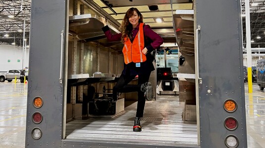 Mahdieh Allahviranloo, an Amazon Visiting Academic, is seen standing in the back of an Amazon delivery vehicle. The truck is empty, it's back door is open, and Mahdieh is smiling while wearing a safety vest.