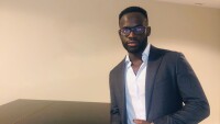 Oritseweyinmi Henry Ajagbawa poses in front of a wall in a suit jacket and a collared shirt, he is leaning on a shiny dark table