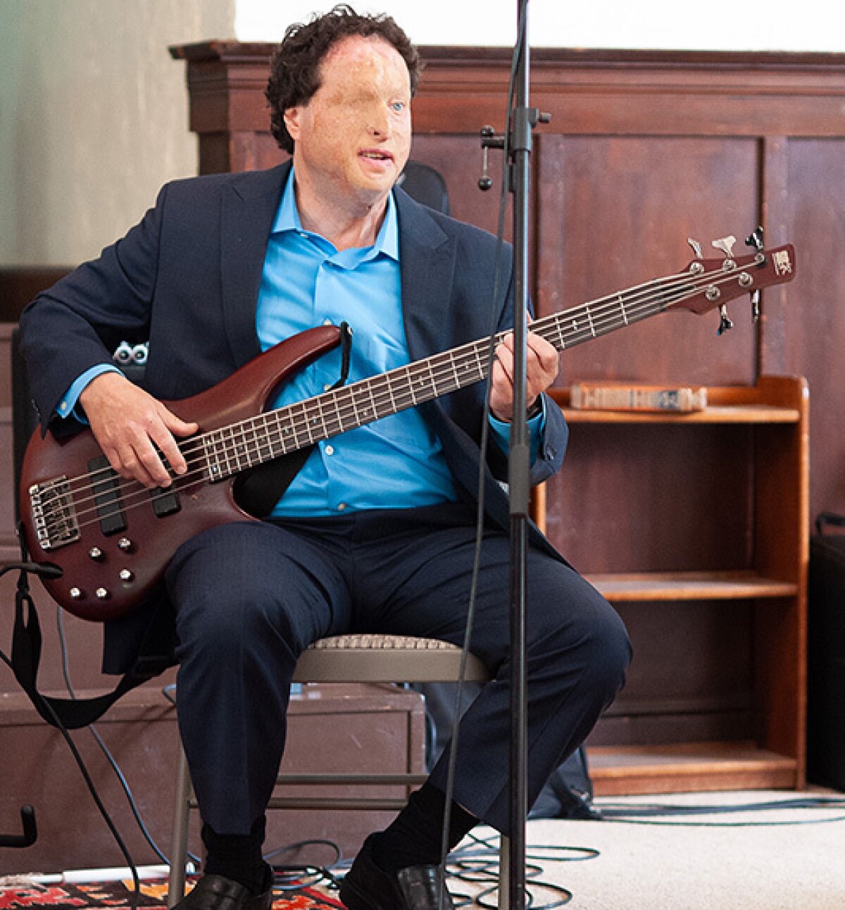Josh Miele is seen wearing a suit, sitting in a chair while playing a bass guitar, there is a mic stand and an amp in the foreground