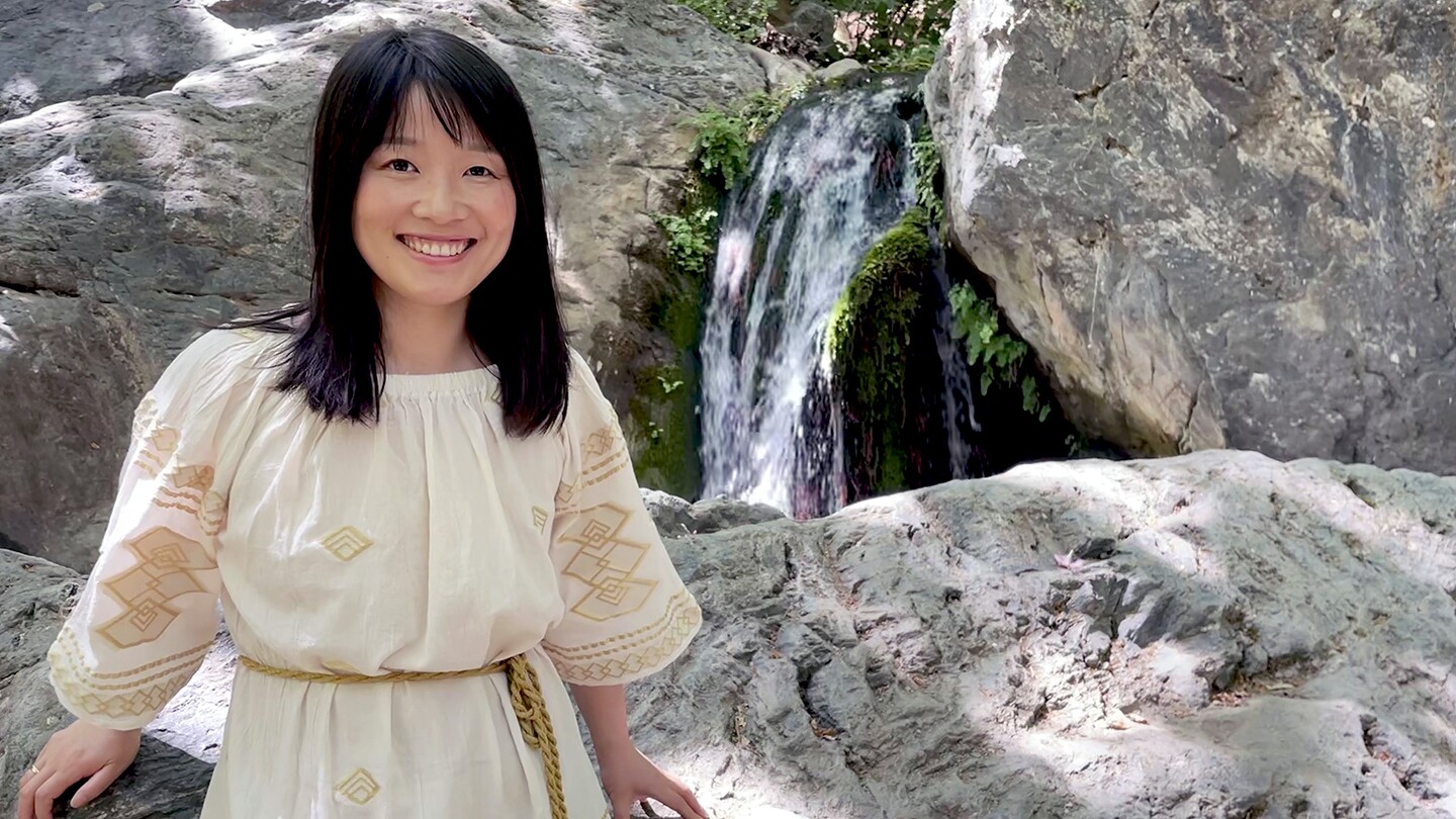 Linghui Luo is seen standing outside in front of a rock formation, with a small waterfall in the background