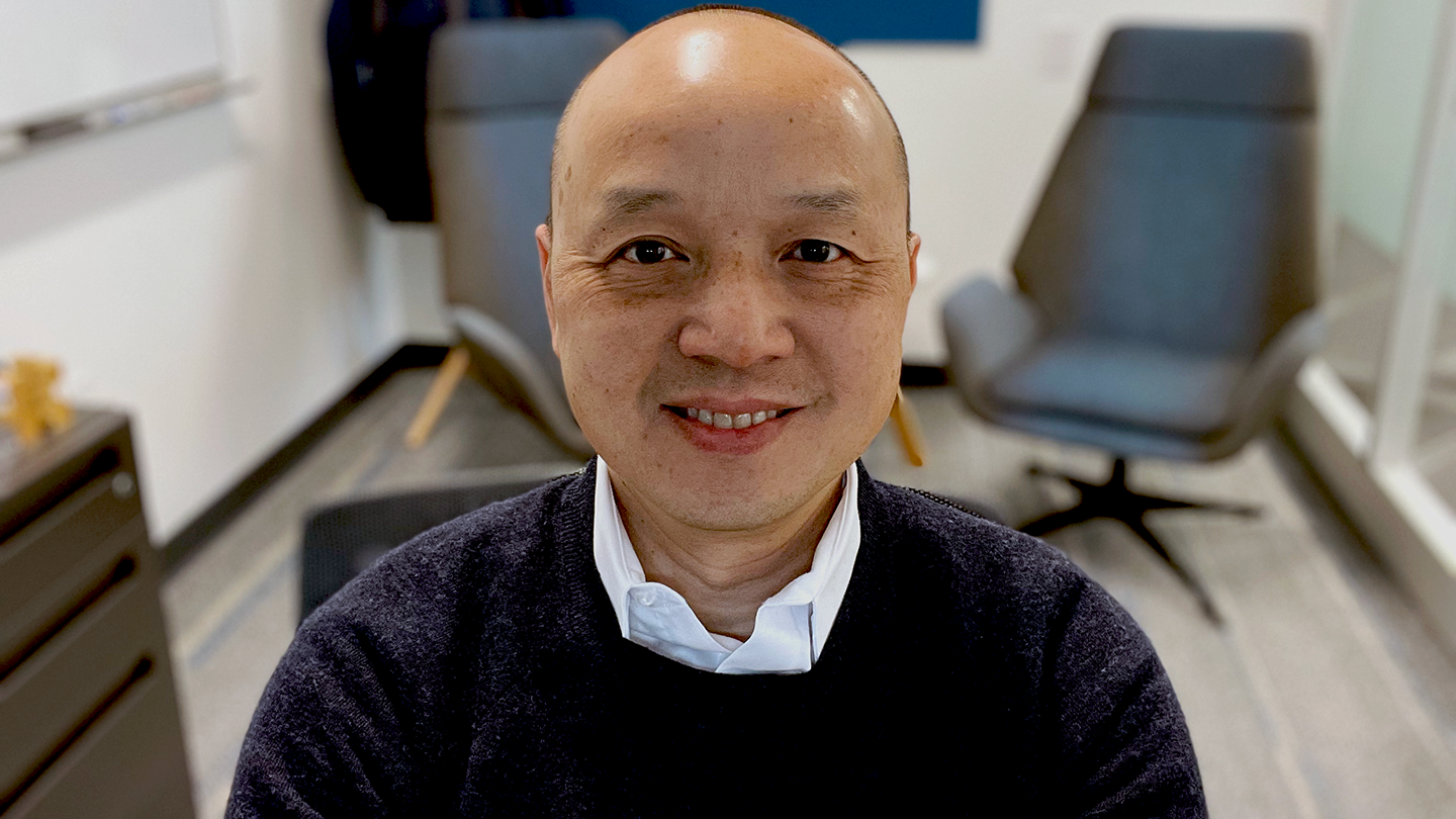 Amazon senior principal engineer Luu Tran is seen sitting indoors, staring into the camera while smiling, he is wearing a sweater over a dress shirt and there are chairs, a desk, and a whiteboard in the background