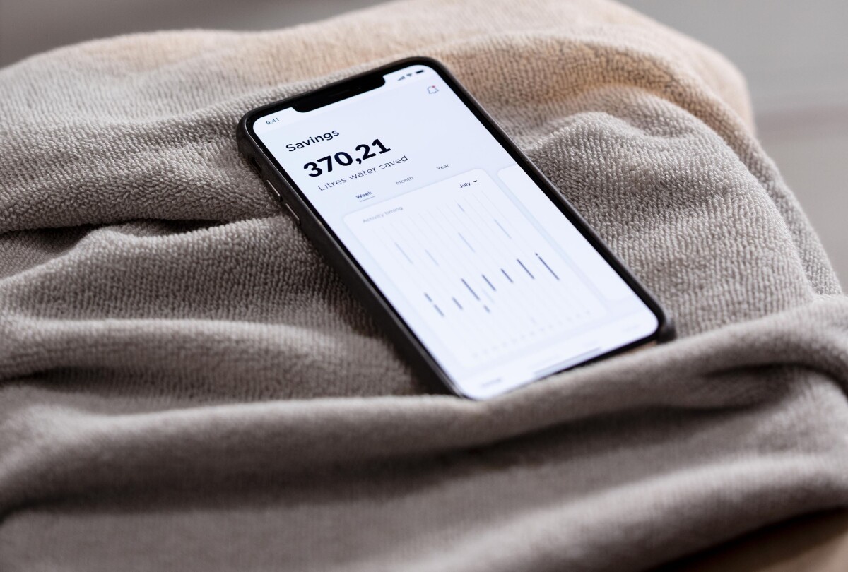 The Orbital Shower mobile app is seen on a smartphone screen displaying 370 liters of water saved, the phone is sitting on a towel