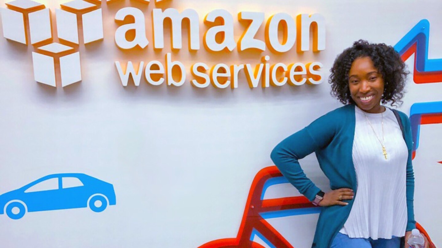 De'Aira Bryant, who has done two internships at Amazon, and is a fourth-year computer science PhD student at the Georgia Institute of Technology, is seen posing in front of a wall with some transportation logos and Amazon Web Services written on it