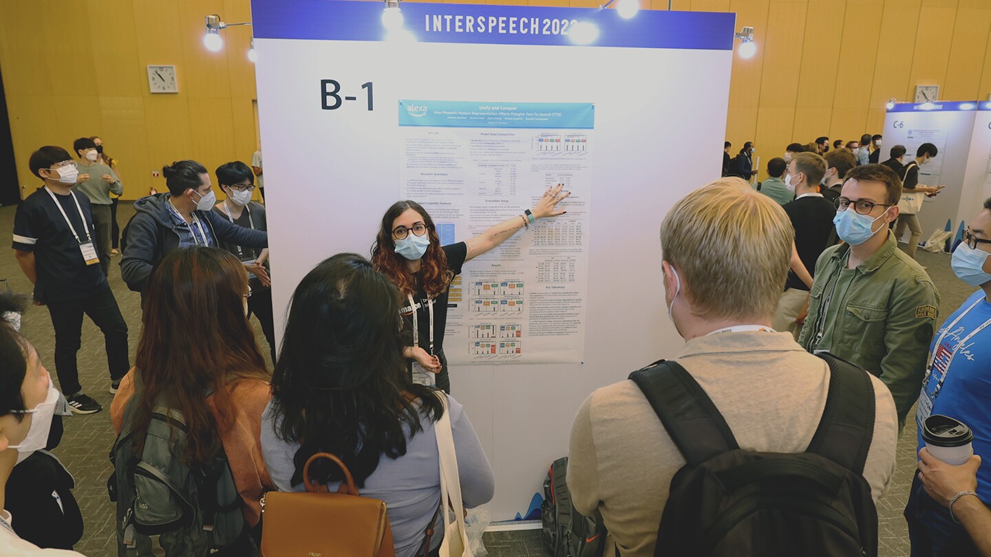 Ariadna Sanchez, a text-to-speech research scientist, is seen presenting to a group at Interspeech 2022, she is pointing to a poster and wearing a mask, as are some of the onlookers