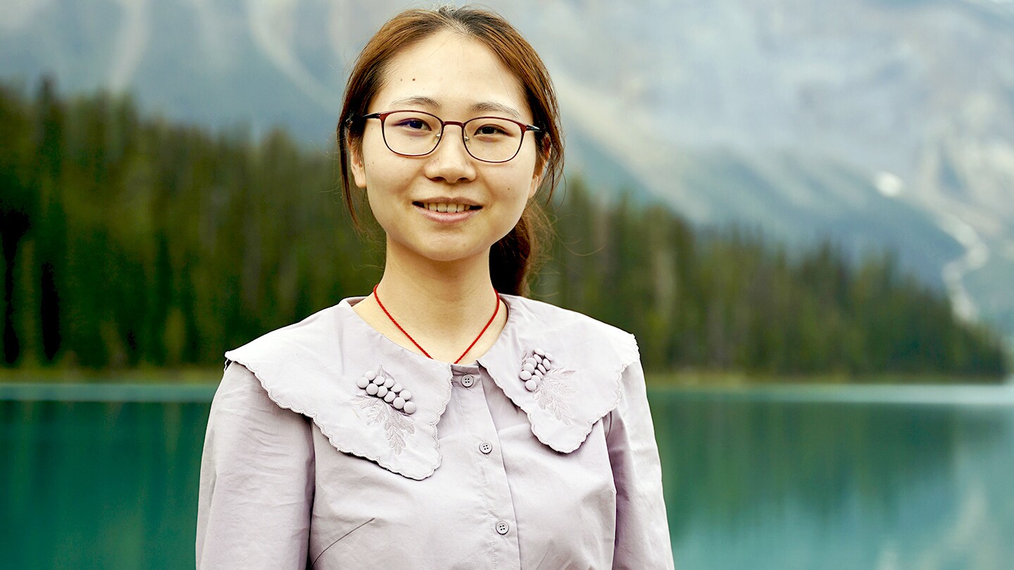 A portrait photo of Qing Guo, she is seen outside with a body of water, trees, and mountains in the background