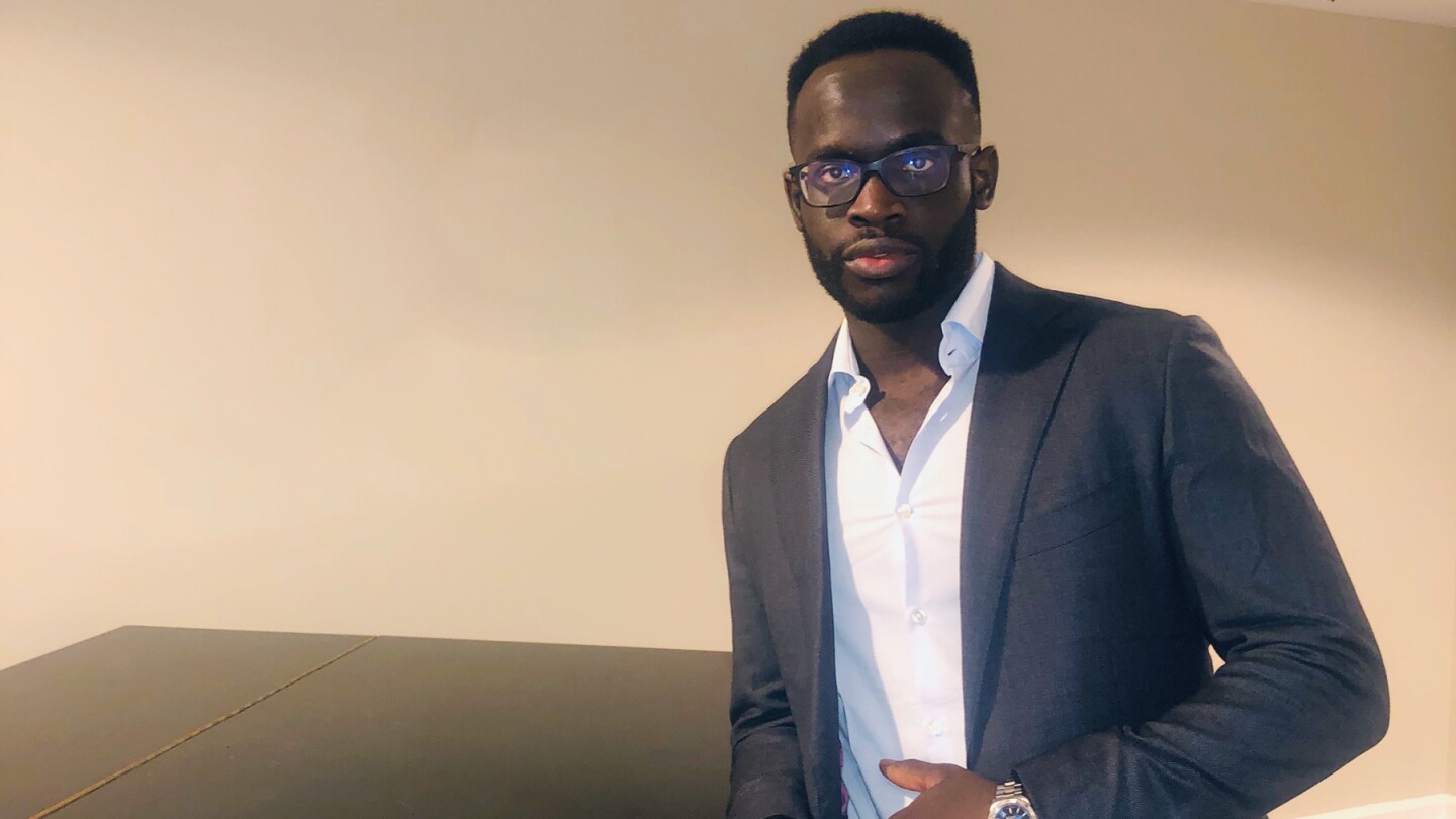 Oritseweyinmi Henry Ajagbawa poses in front of a wall in a suit jacket and a collared shirt, he is leaning on a shiny dark table