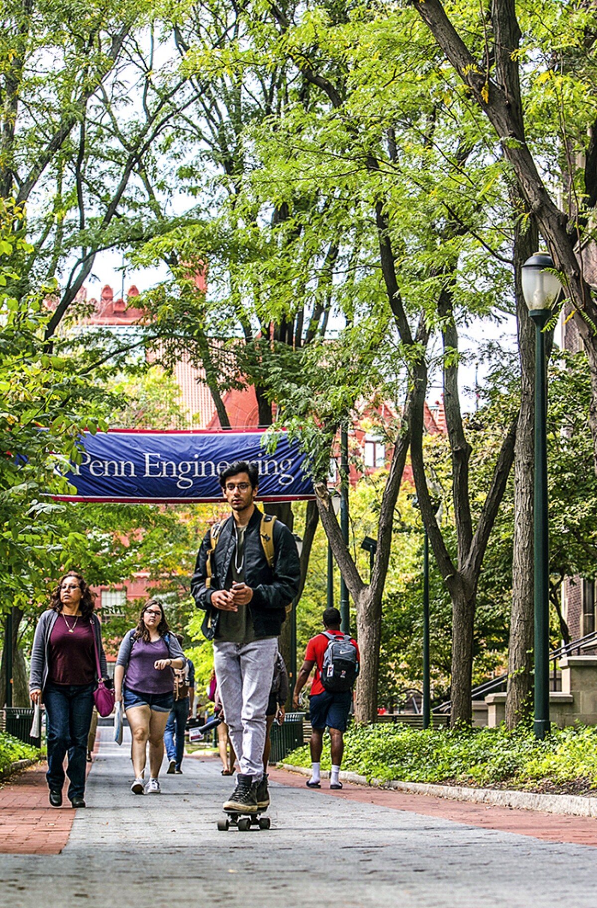 A student is seen skateboarding toward the camera on the Penn Engineering campus, there are students walking behind him on a tree lined walk and there is a Penn Engineering banner hanging over the walkway