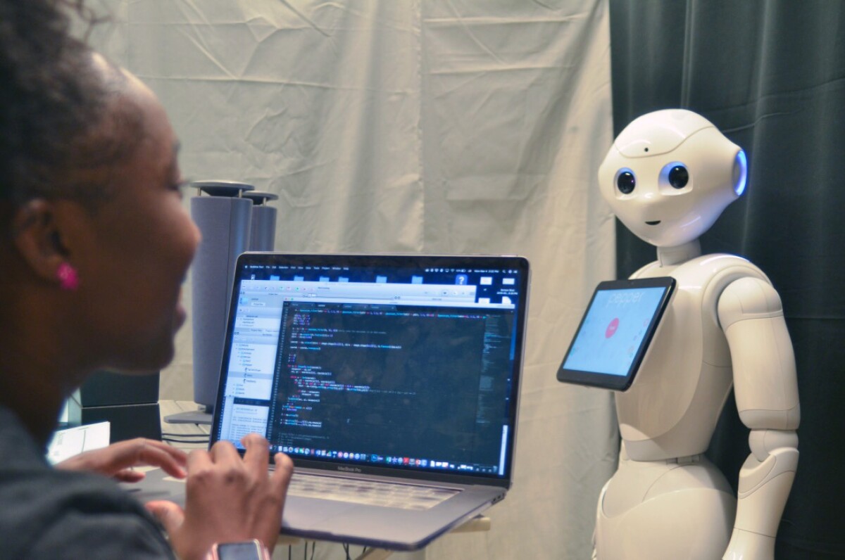 De'Aira Bryant is seen from behind, she is typing on an open laptop and there is a humanoid robot with a display tablet on its chest looking at her to the right of the laptop