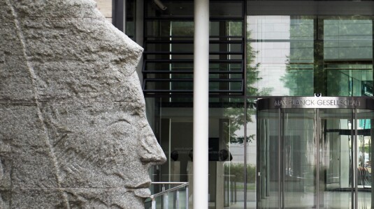 A large stone sculpture is seen outside the entrance to one of the Max Planck Society locations in Germany, a revolving door with the group's name in German is seen to the right