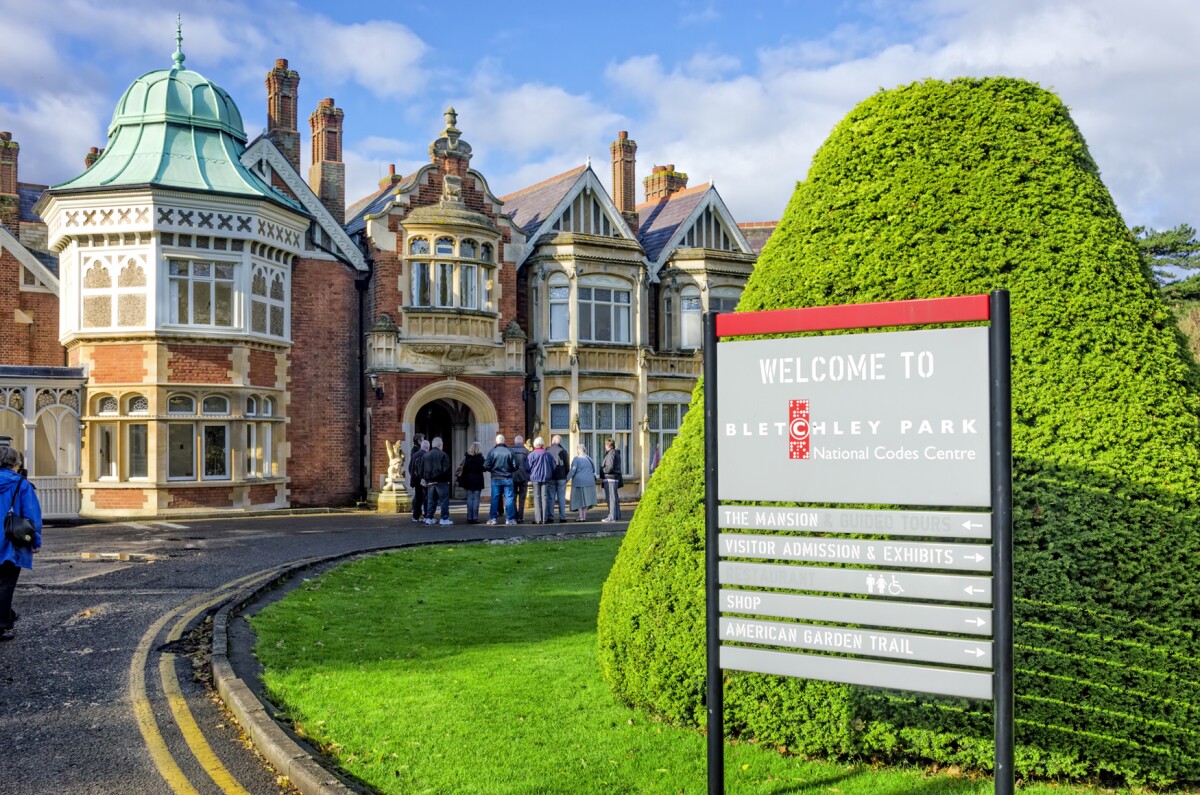 In the foreground, a welcome to Bletchley Park offers a guide, in the background a group of tourists get a guided tour. This area was used in World War 2 to break the German Enigma Codes.