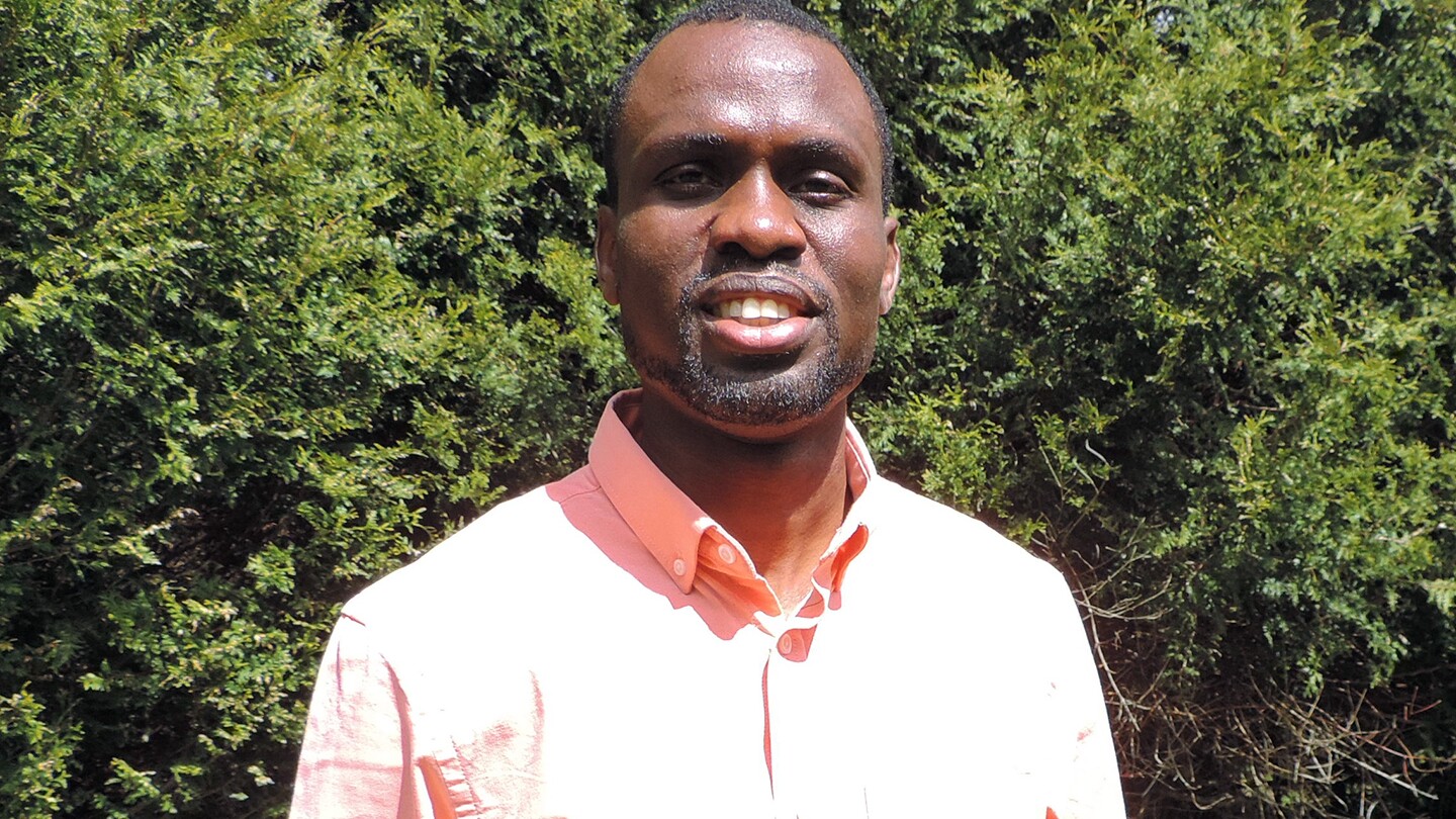 Chukwudi Chukwudozie is seen looking into the camera, smiling while wearing a collared shirt and standing in front of some greenery