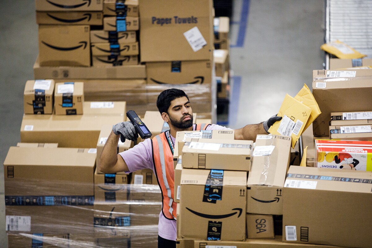 An Amazon employee is seen scanning packages, there are pallets of boxes behind him