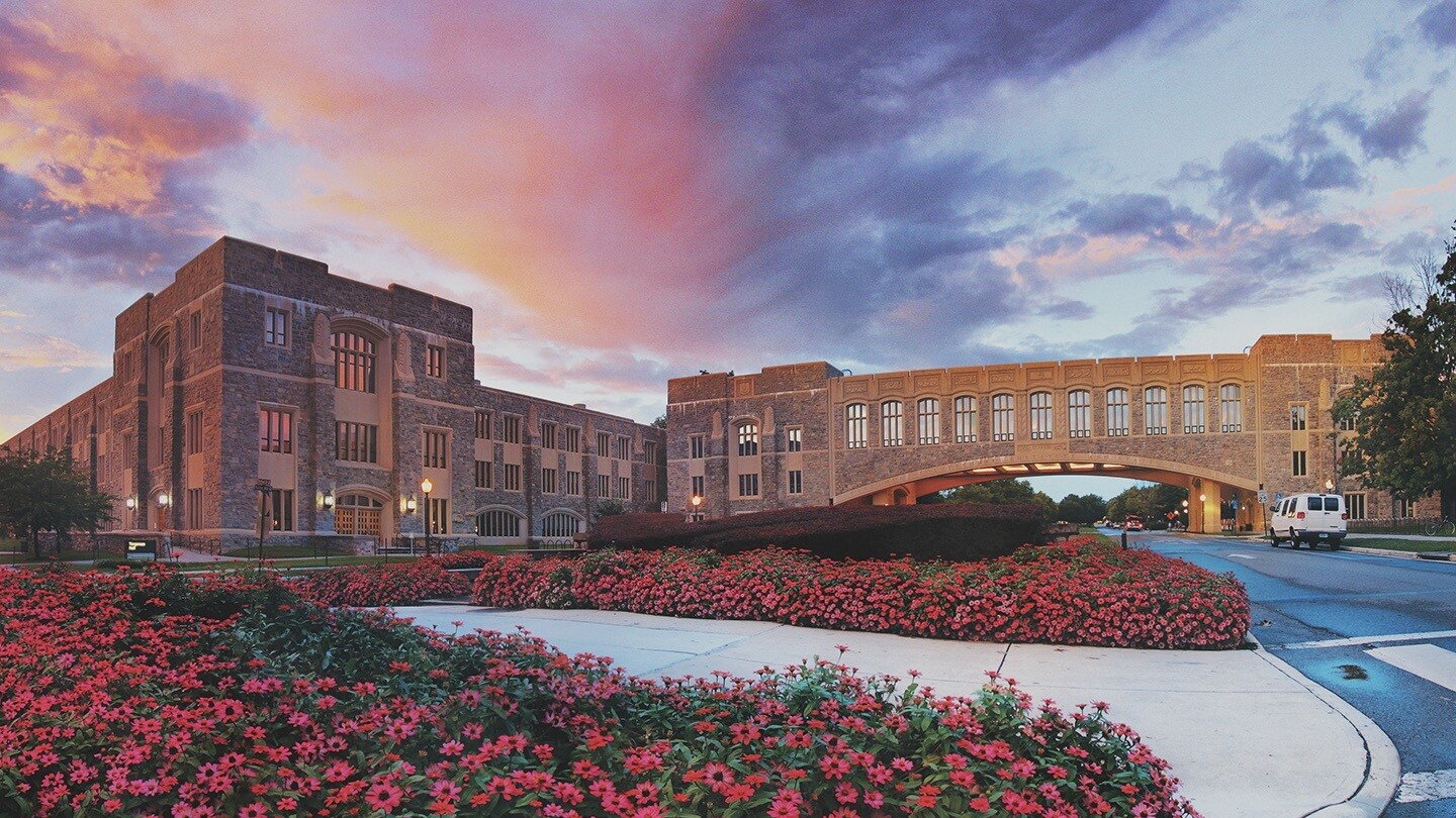 Image shows Torgersen Hall on the campus of Virginia Tech, the building and pedestrian bridge are in the background, flowers are in the foreground, the sky is streaked with clouds