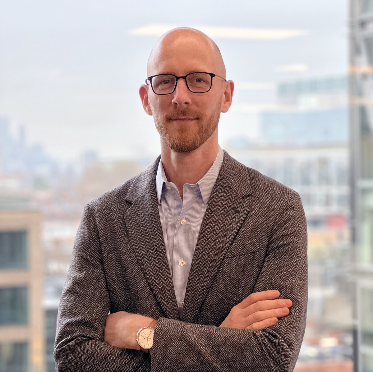 Thomas Hoe, principal economist, is seen standing in front of a widow in a blazer and dress shirt, his arms are crossed and there is a cityscape visible in the backgound.