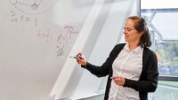 Éva Tardos, the Jacob Gould Schurman Professor of Computer Science at Cornell University, is seen standing at a whiteboard with an equation written on it, holding a marker in her hand