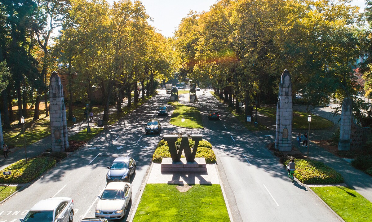 An aerial shot shows cars entering and exiting the University of Washington campus via a tree lined route on a sunny day, a median divides the lanes and a large W sits in the center of the median.
