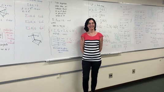 Luciana Buriol is seen standing in front of a large whiteboard filled with equations, she is standing toward the center, smiling with her hands clasped behind her back