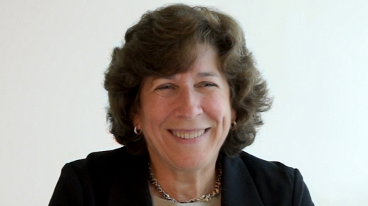 Portrait of McKeown from the waist up, in a business suit, seated at a desk with her hands clasped in front of her.