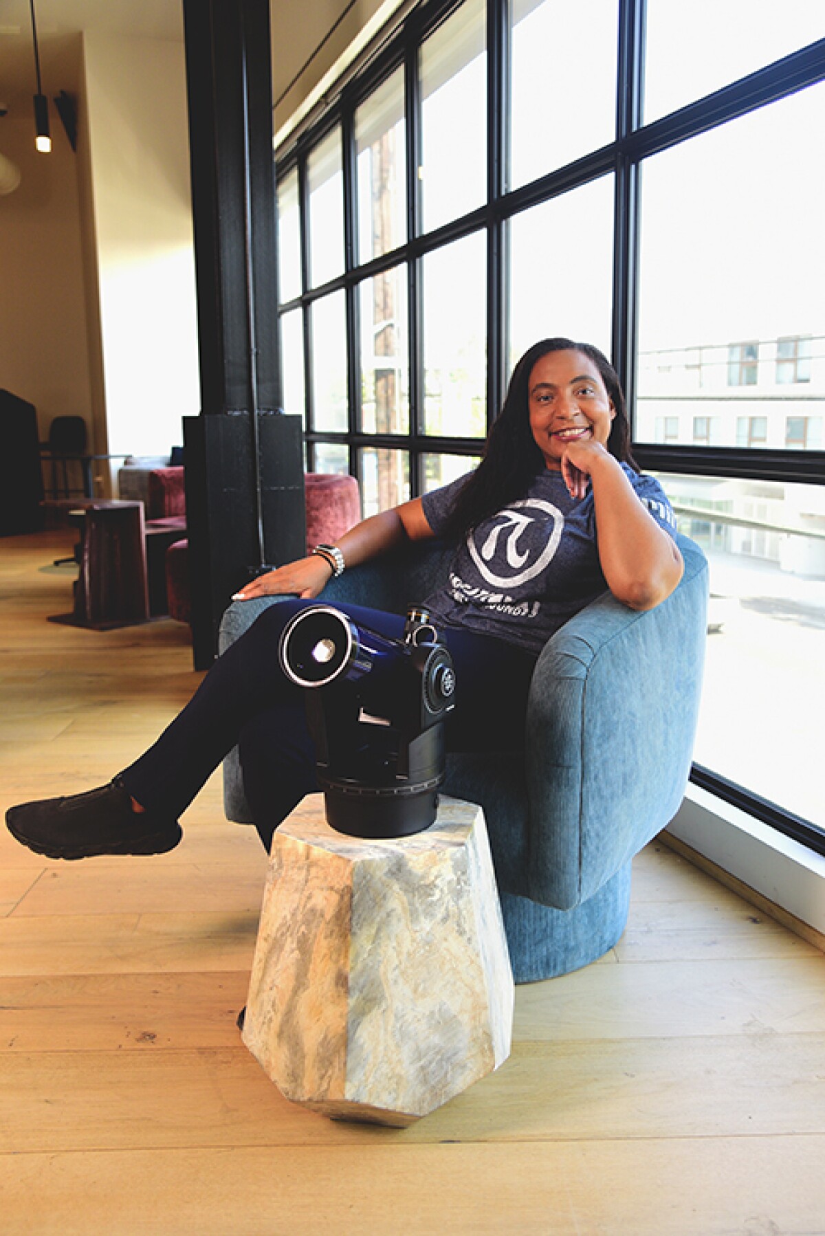 Nia Jetter is seen sitting in a chair with a telescope on a stand in front of her and windows behind her, she is smiling into the camera