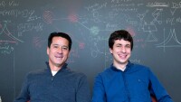 Michael Kearns and Aaron Roth seated at a table in front of a large chalk board.