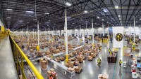 This picture is an overhead shot inside an Amazon center, workers can be seen moving amidst hundreds of boxes which sit on conveyor belts and carts, in the upper left foreground, a yellow railing extends into the distance.