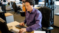 Josh Miele, in a purple dress shirt, sits at a desk in an office, he is typing and looking at a computer screen, there are chairs and desks in the background