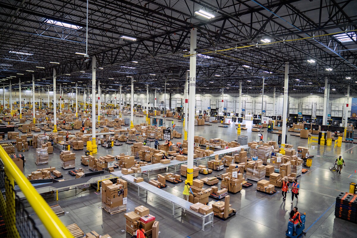 An overhead shot inside an Amazon fulfillment center shows hundreds of boxes on conveyor belts along with people monitoring the flow of those packages