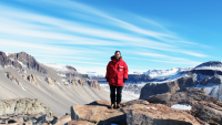 Jonathan Toner is seen in Antartica, he is standing on some large rocks wearing a red parka with a mountain range in the background