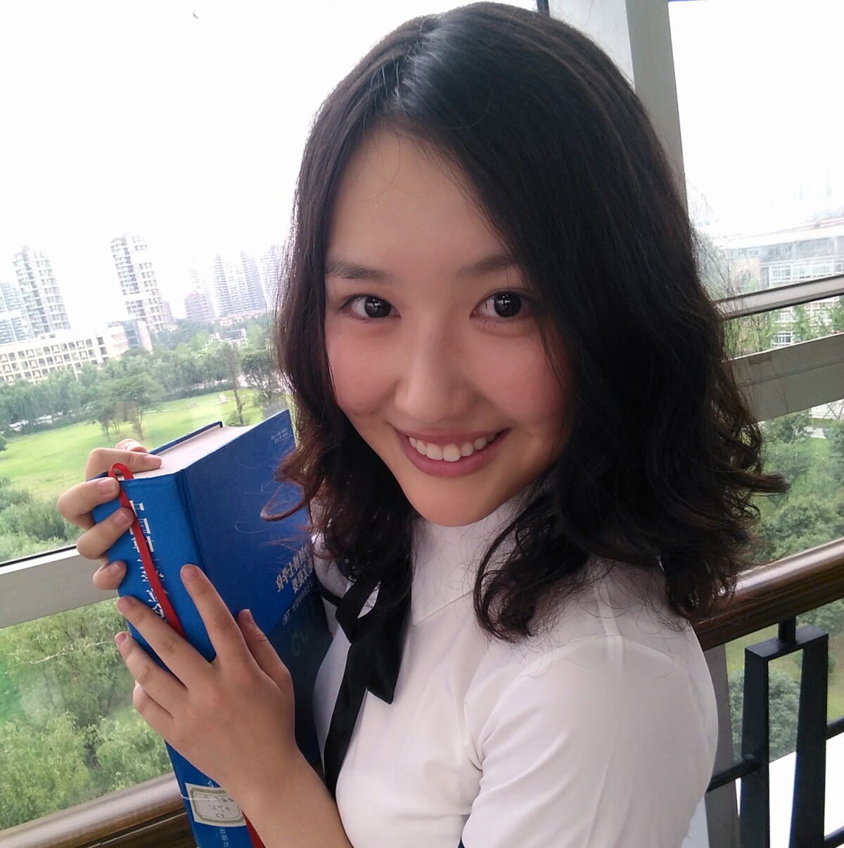 Minghui He, a research scientist, is seen smiling and holding a book with a park and cityscape in the background