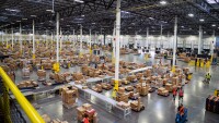 An overhead shot inside an Amazon fulfillment center shows hundreds of boxes on conveyor belts along with people monitoring the flow of those packages