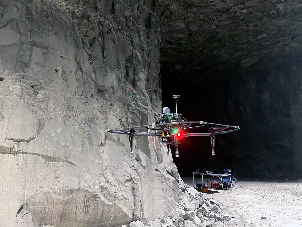 Image shows a drone hovering in the air inside of a mine in the foreground, there is a table with some equipment on it and a chair in the background