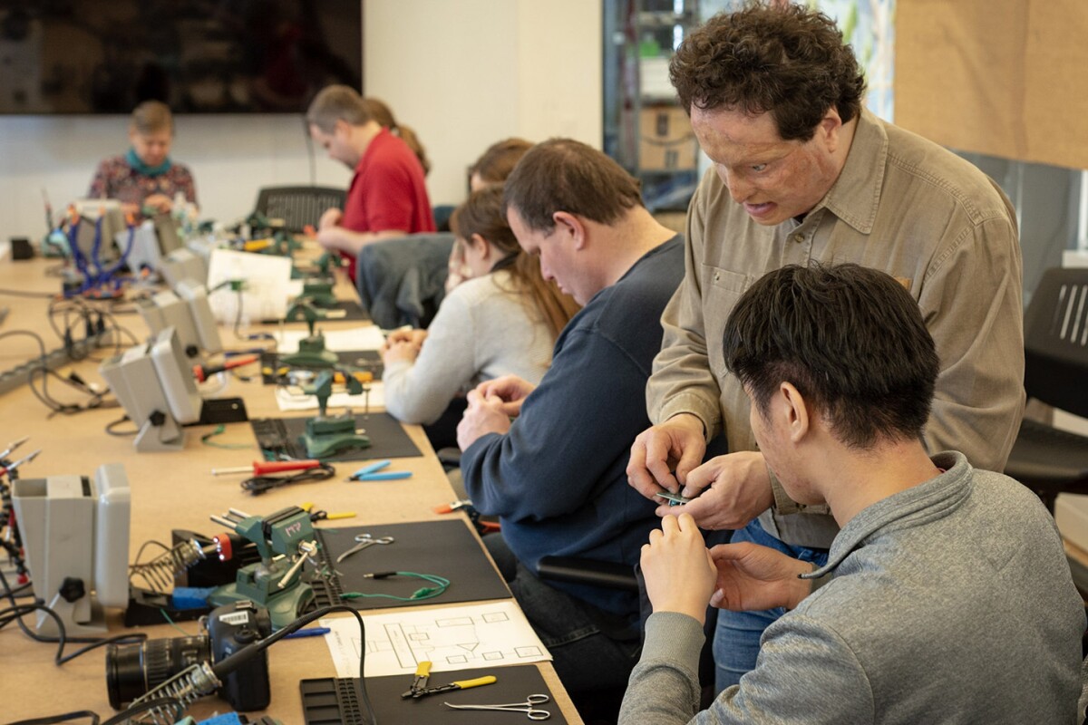 Josh Miele, standing, gives advice to a student who is seated during a soldering workshop. Several students are sitting at a large table with soldering equipment.
