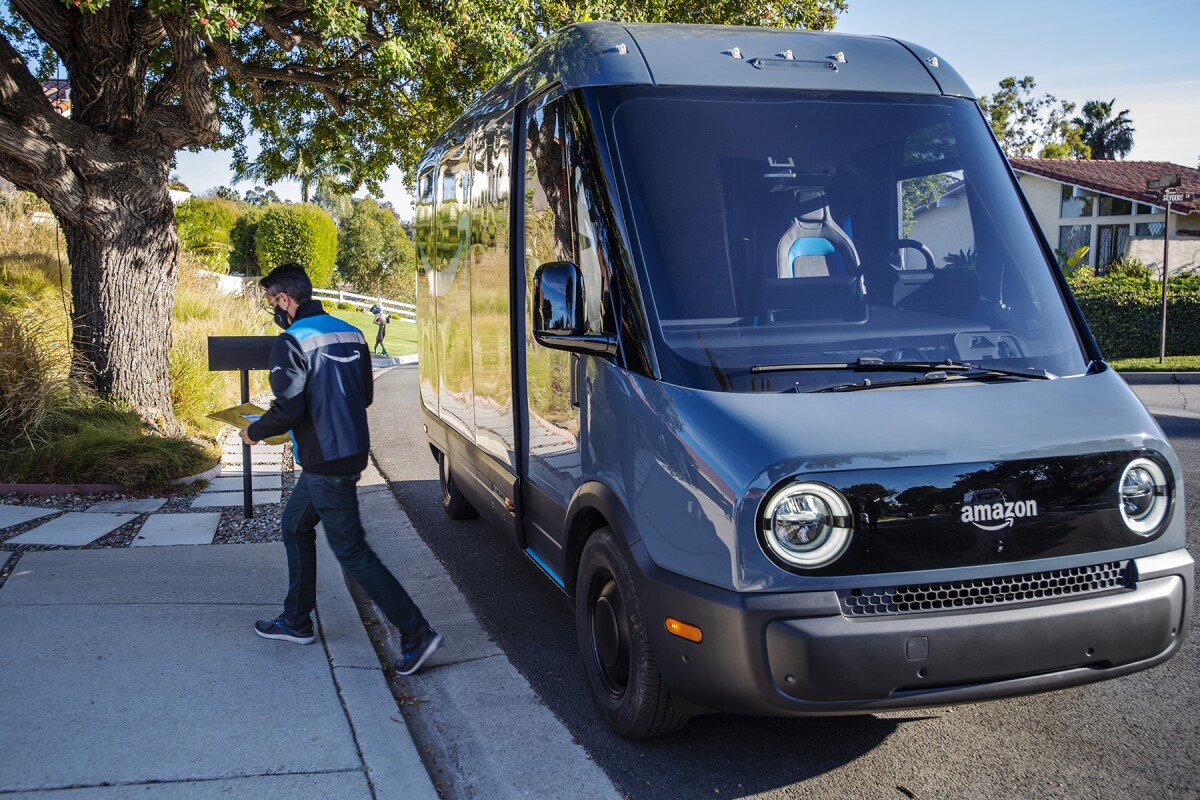 An Amazon employee is seen making a delivery while an electric delivery van is parked behind him on a residential street in Los Angeles