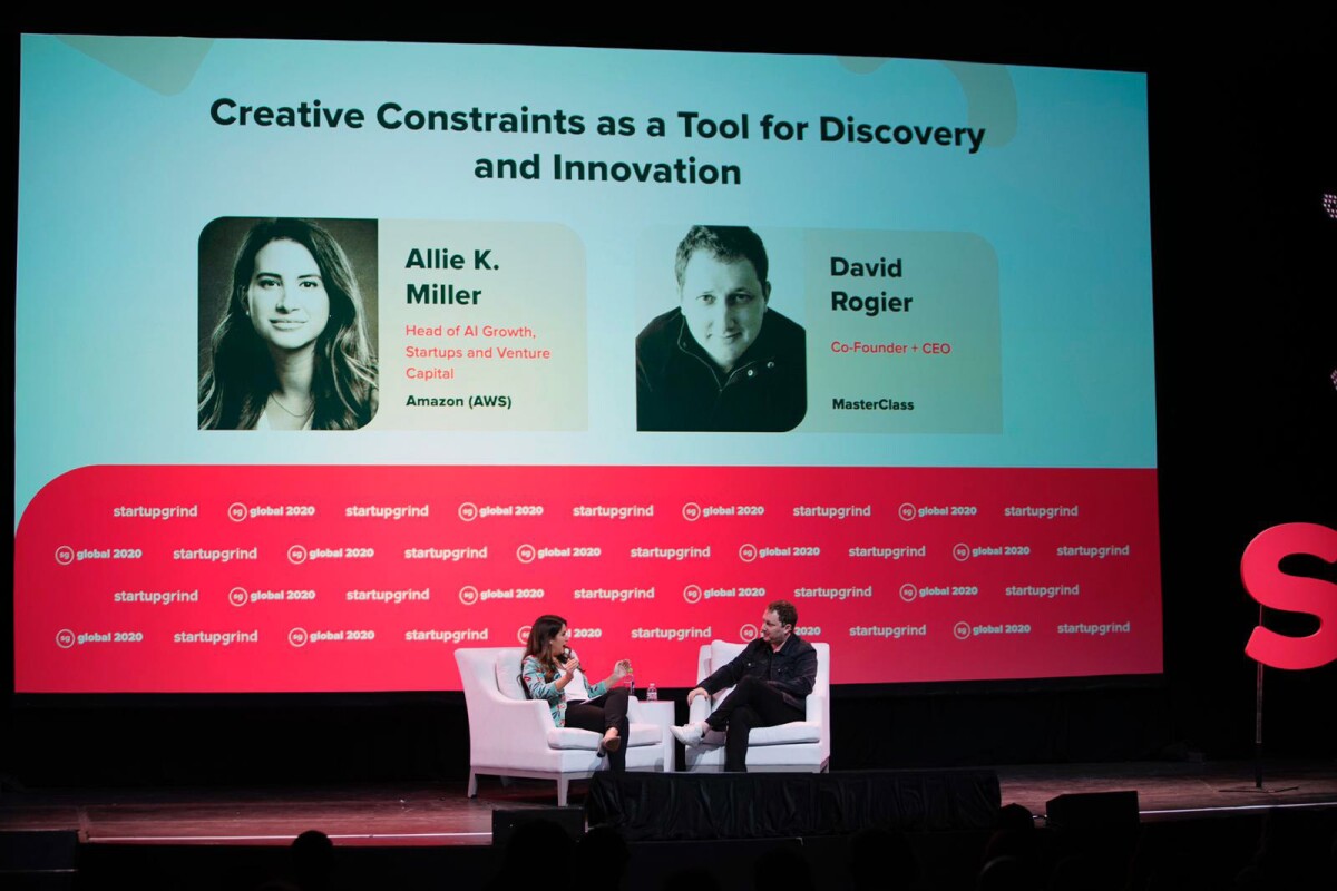 Allie Miller, left, chats with with David Rogier, the CEO and co-founder of MasterClass, right, on stage at an event. Both are seated in white chairs, behind them is a large display screen with the title of their talk, their names, pictures, and titles.