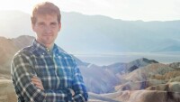 Sergey Menis is seen outside on a sunny day with a colorful landscape of mountains behind him, Sergey is standing to the left with his arms crossed, looking into the camera