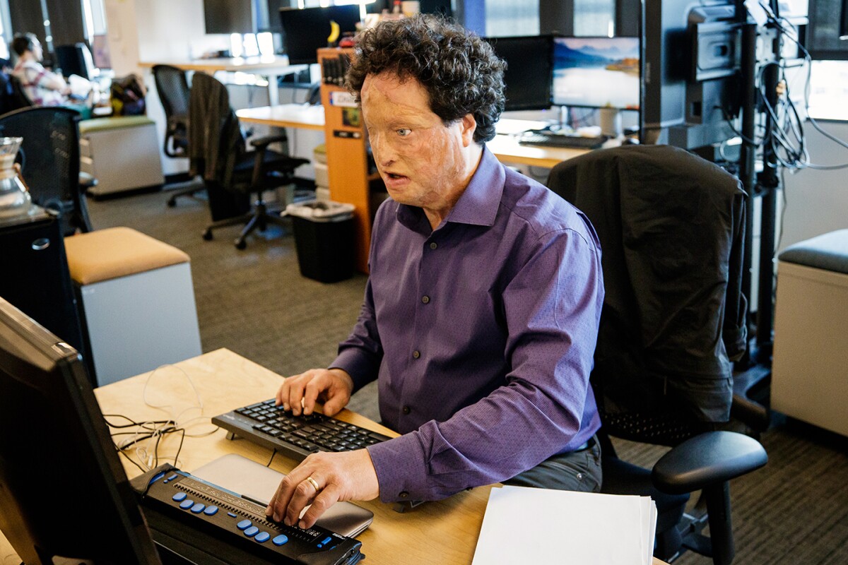 Josh Miele, in a purple dress shirt, sits at a desk in an office, he is typing and looking at a computer screen, there are chairs and desks in the background