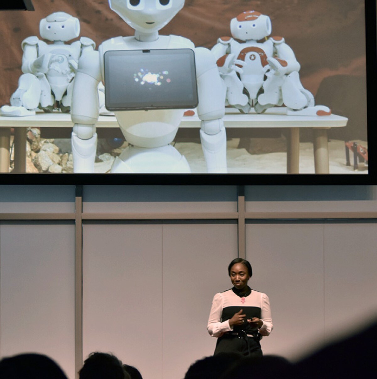 De'Aira Bryant is seen standing on a stage with a screen elevated above her in the background showing robots at her TEDx Talk