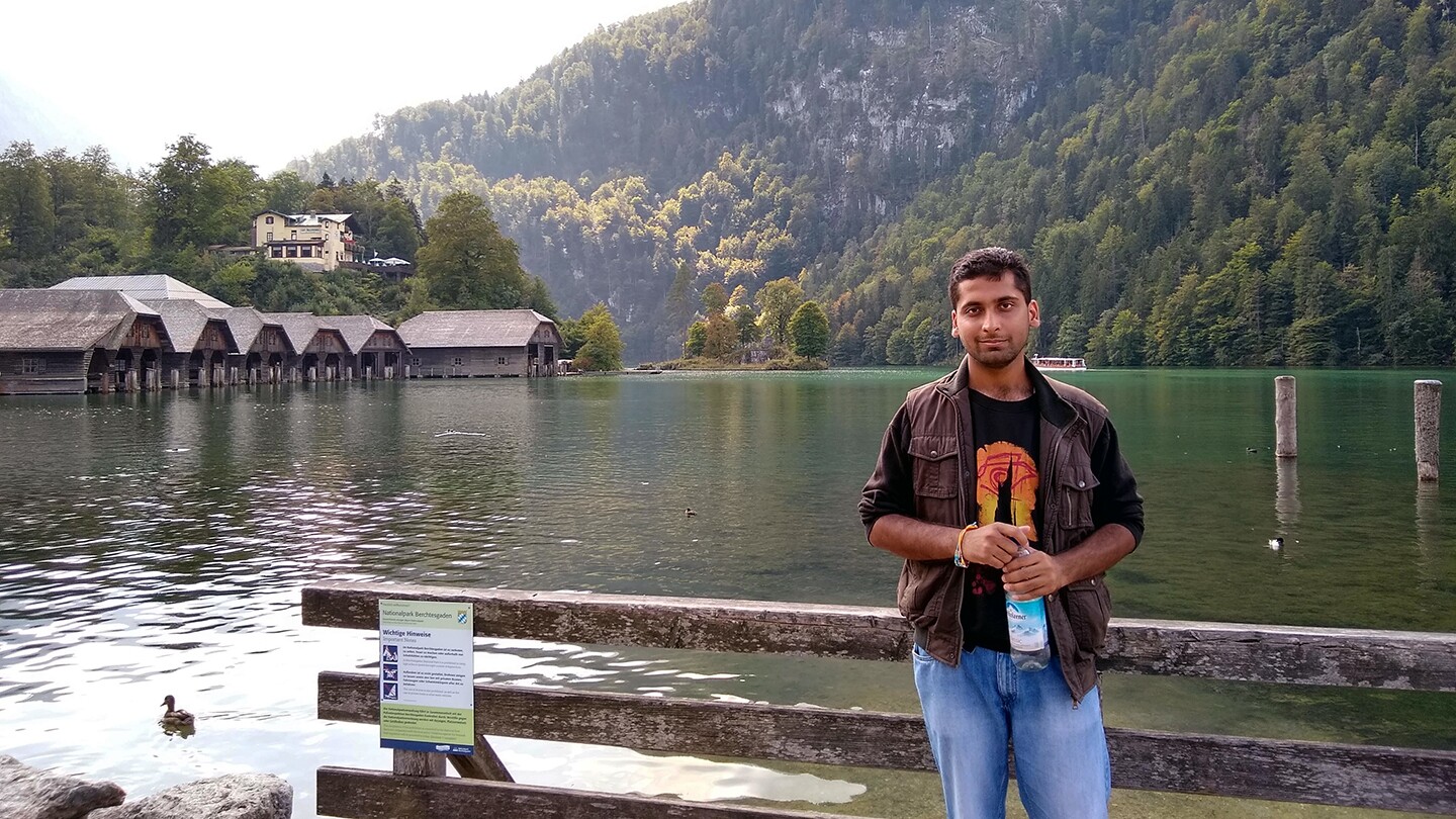 Ankan Bansal, an applied scientist at Amazon, is seen standing in front of a large body of water on a bright day with tree covered mountains in the background